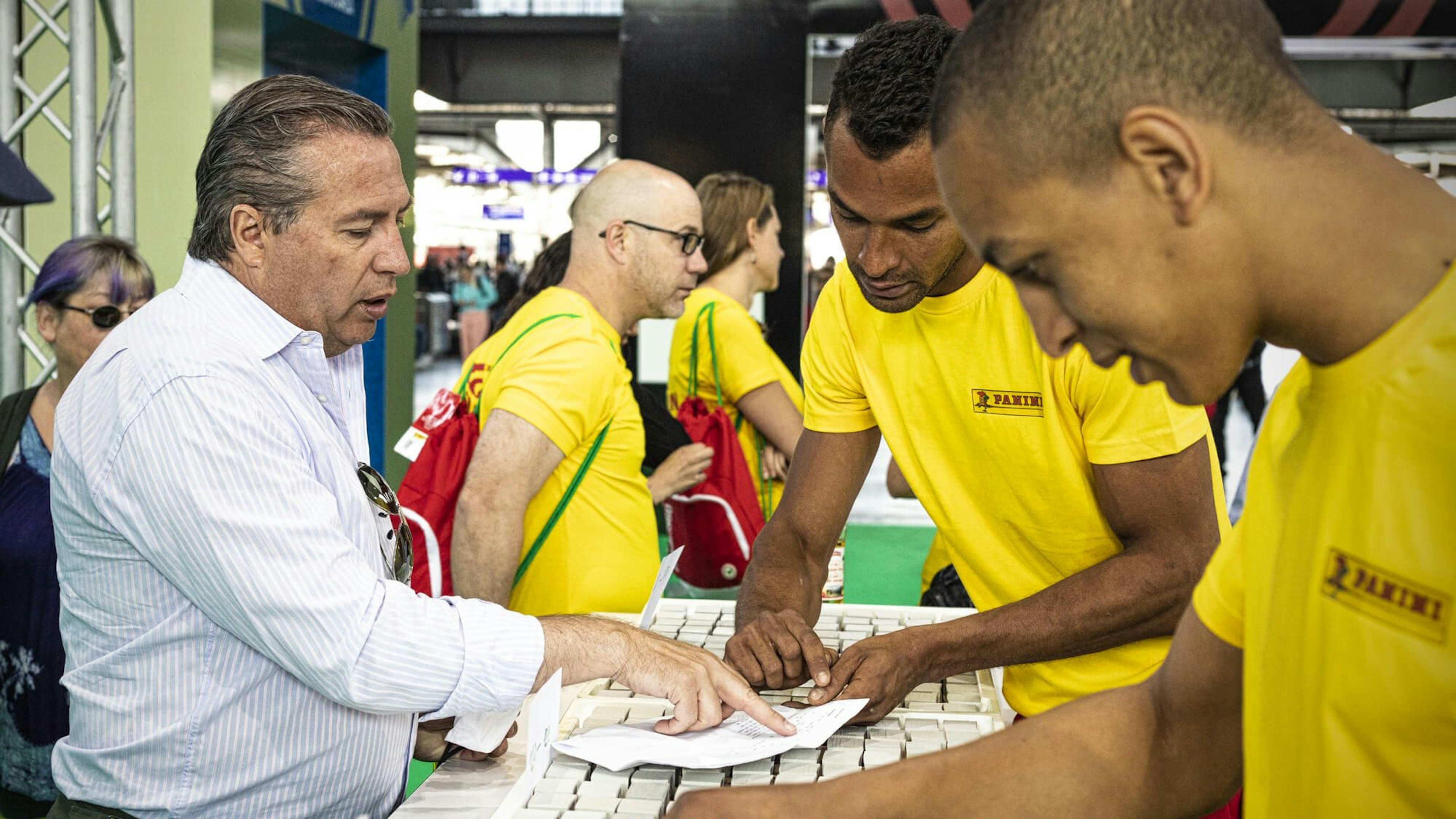 A man interacts with Panini staff members wearing yellow shirts at the Panini Stadion event, discussing or examining items on a table. The setting appears to be busy with other participants in the background.