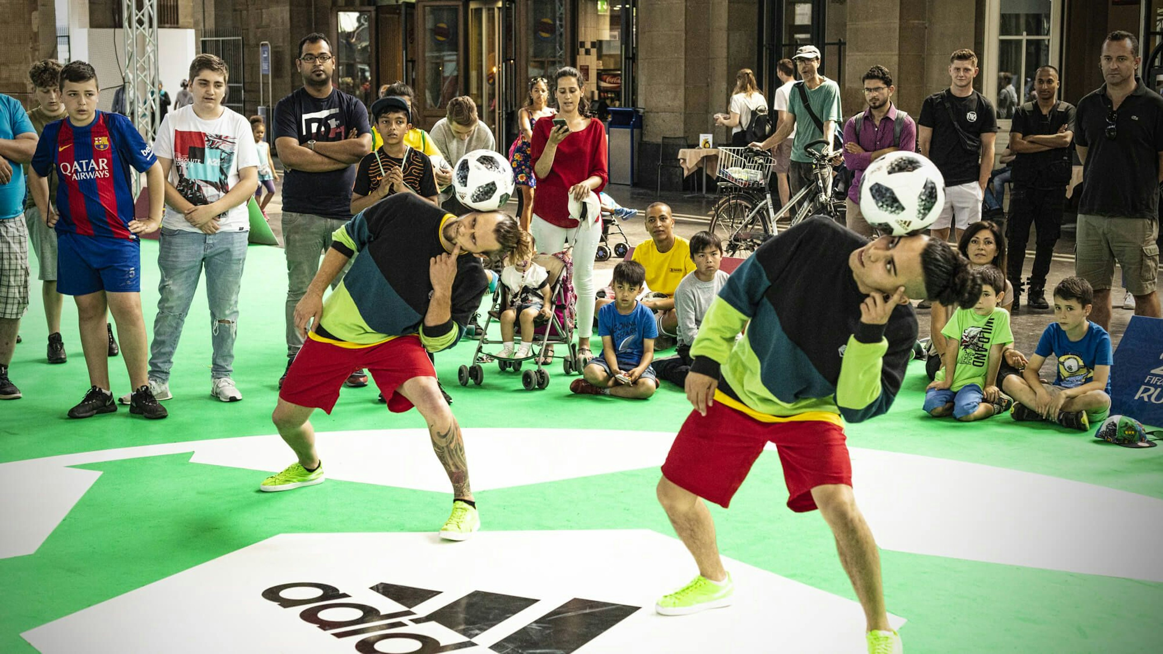 Two freestyle footballers perform tricks with soccer balls on a green Adidas-branded field, watched by an attentive crowd.