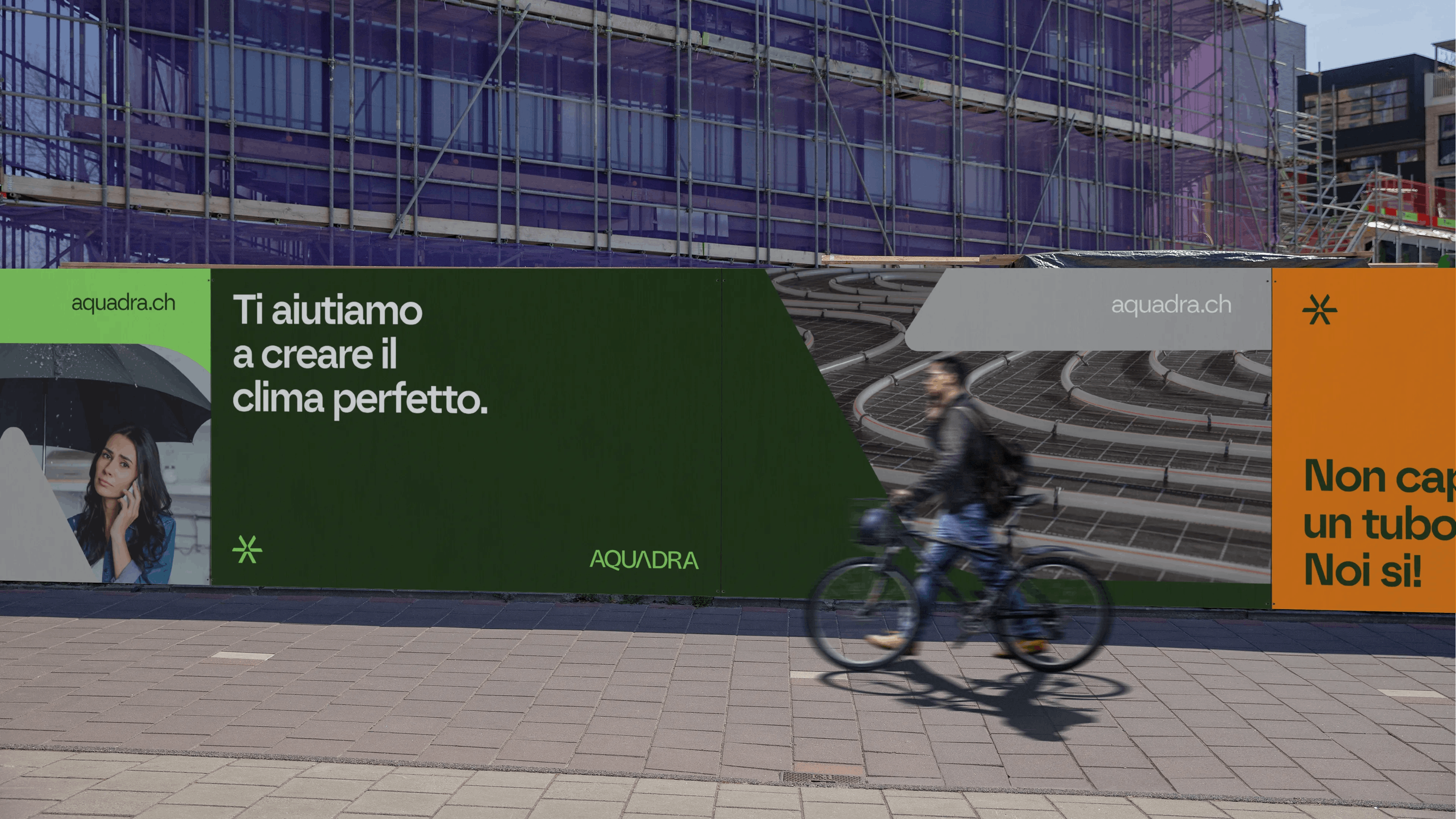 A cyclist rides past a construction site billboard featuring the company 'AQUADRA' with the text 'Ti aiutiamo a creare il clima perfetto' and another section with the phrase 'Non capisci un tubo? Noi si!