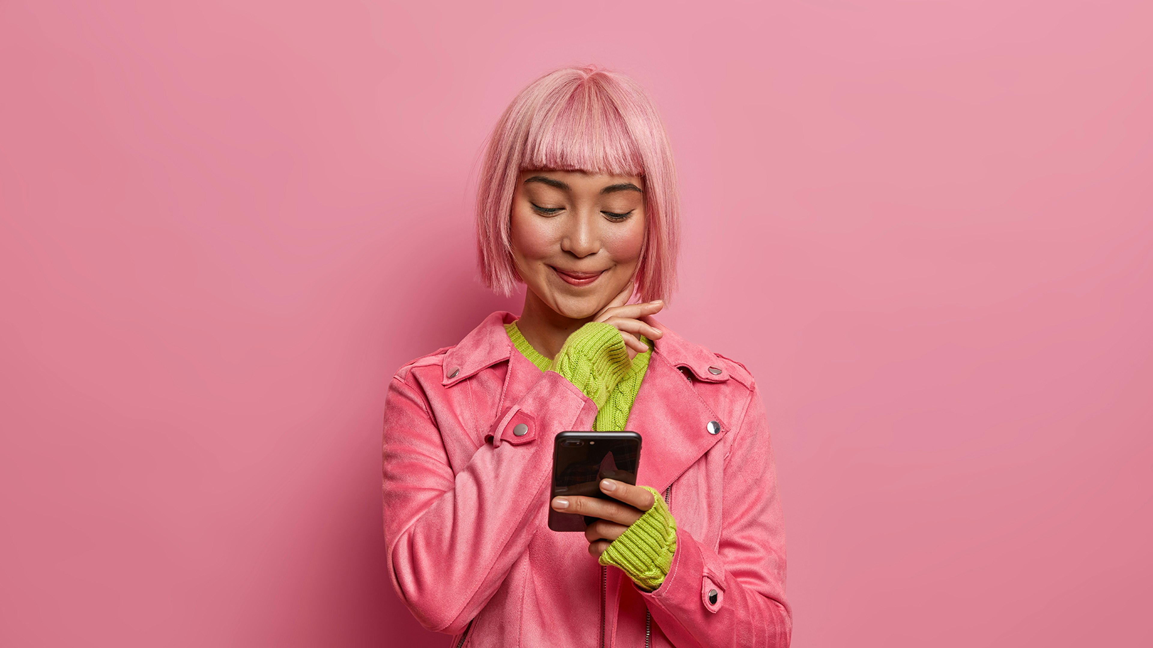 ragazza che guarda lo smartphone con sguardo attento e felice, capelli e felpa e background rosa
