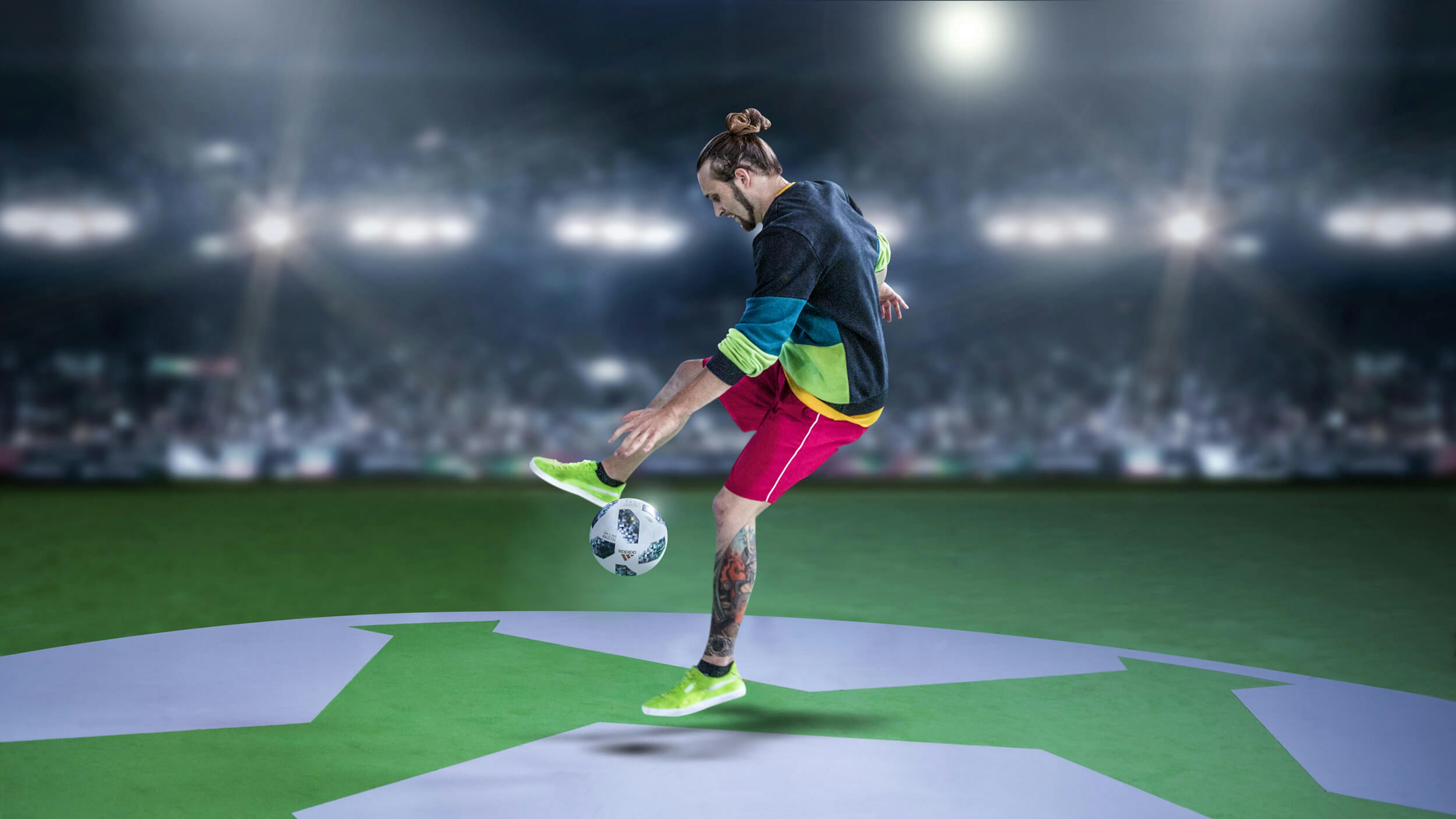 A freestyle footballer performs a trick with a soccer ball in a well-lit stadium, standing on a green field with an Adidas logo. The background shows a blurred crowd, emphasizing the dynamic action.