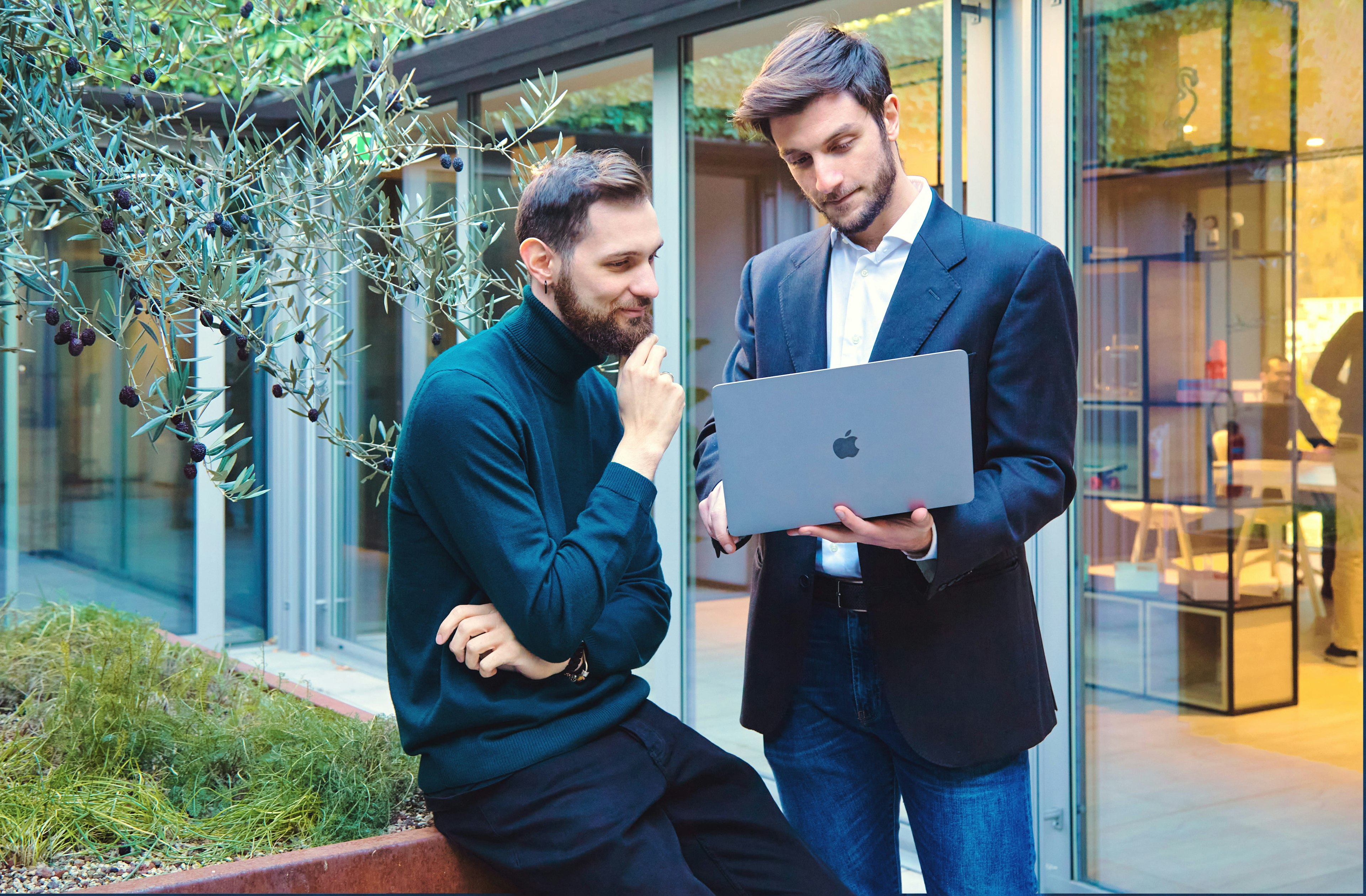 Two men discussing work on a laptop in an outdoor setting with a modern office backdrop, engaged in a thoughtful conversation.