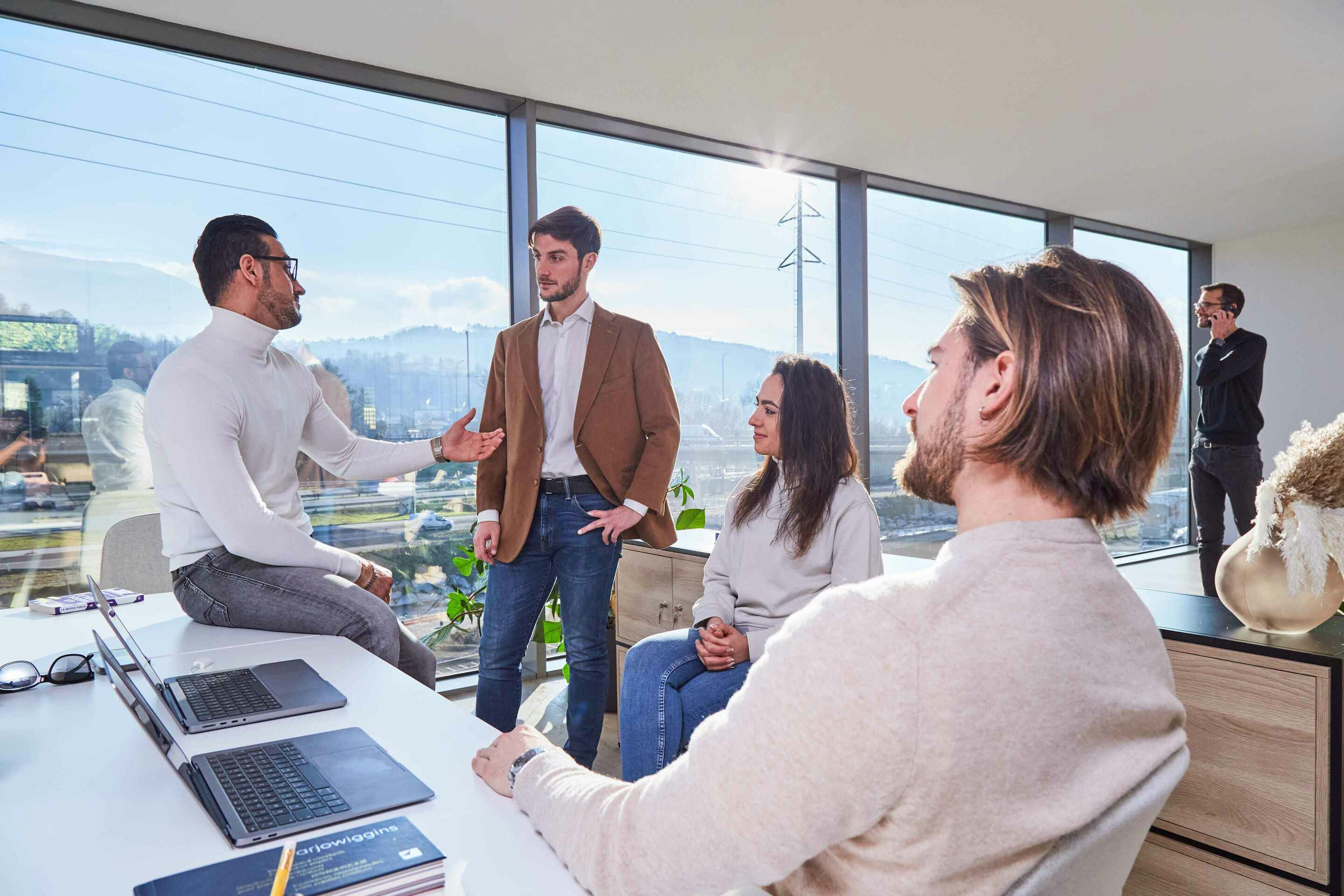 Five professionals engaged in a discussion in a modern, well-lit office with a scenic view, using laptops and actively communicating.