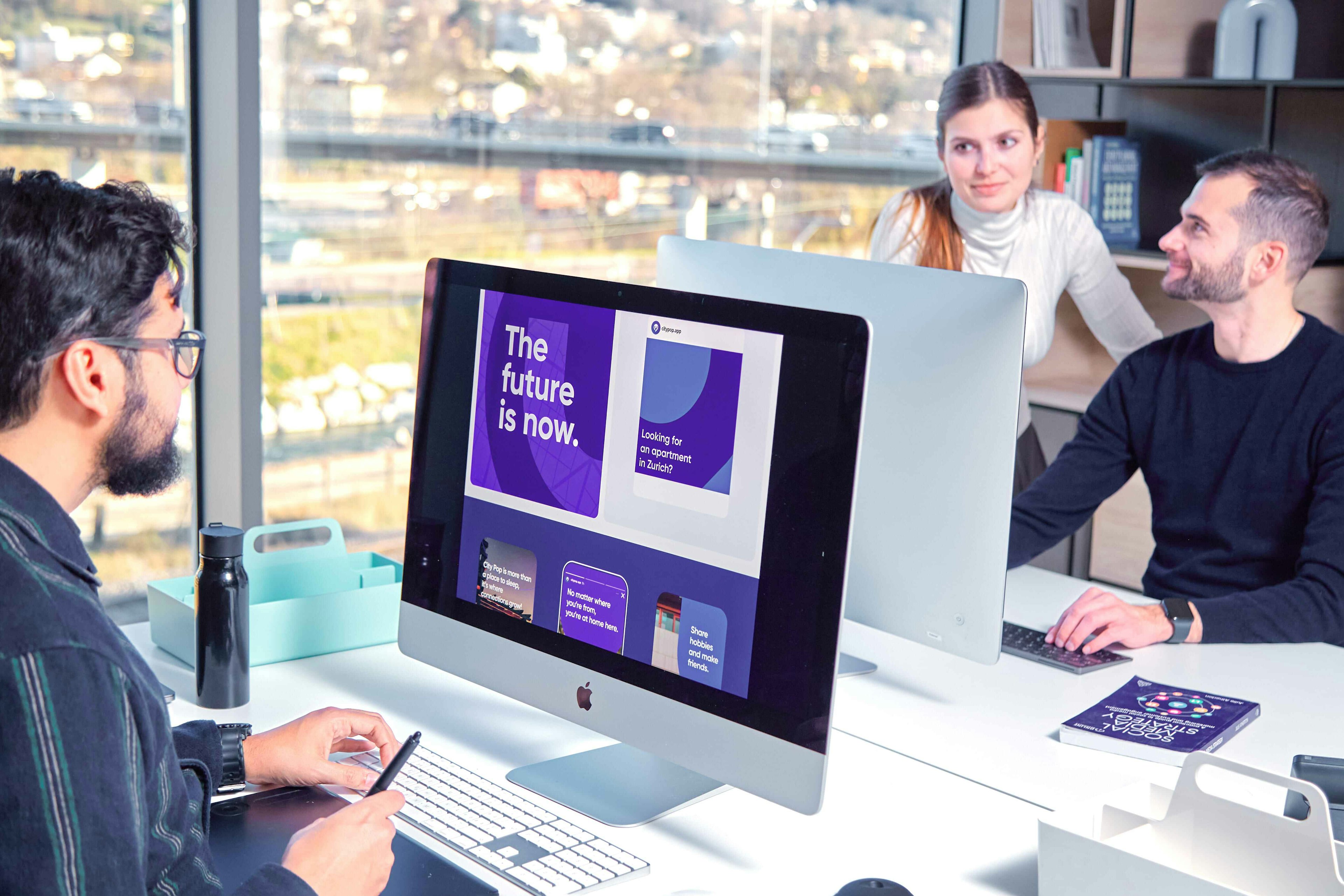 Three professionals in a modern office setting, collaborating around a large iMac displaying a vibrant purple presentation with the text 'The future is now.' The environment offers a view of an urban landscape through large windows.