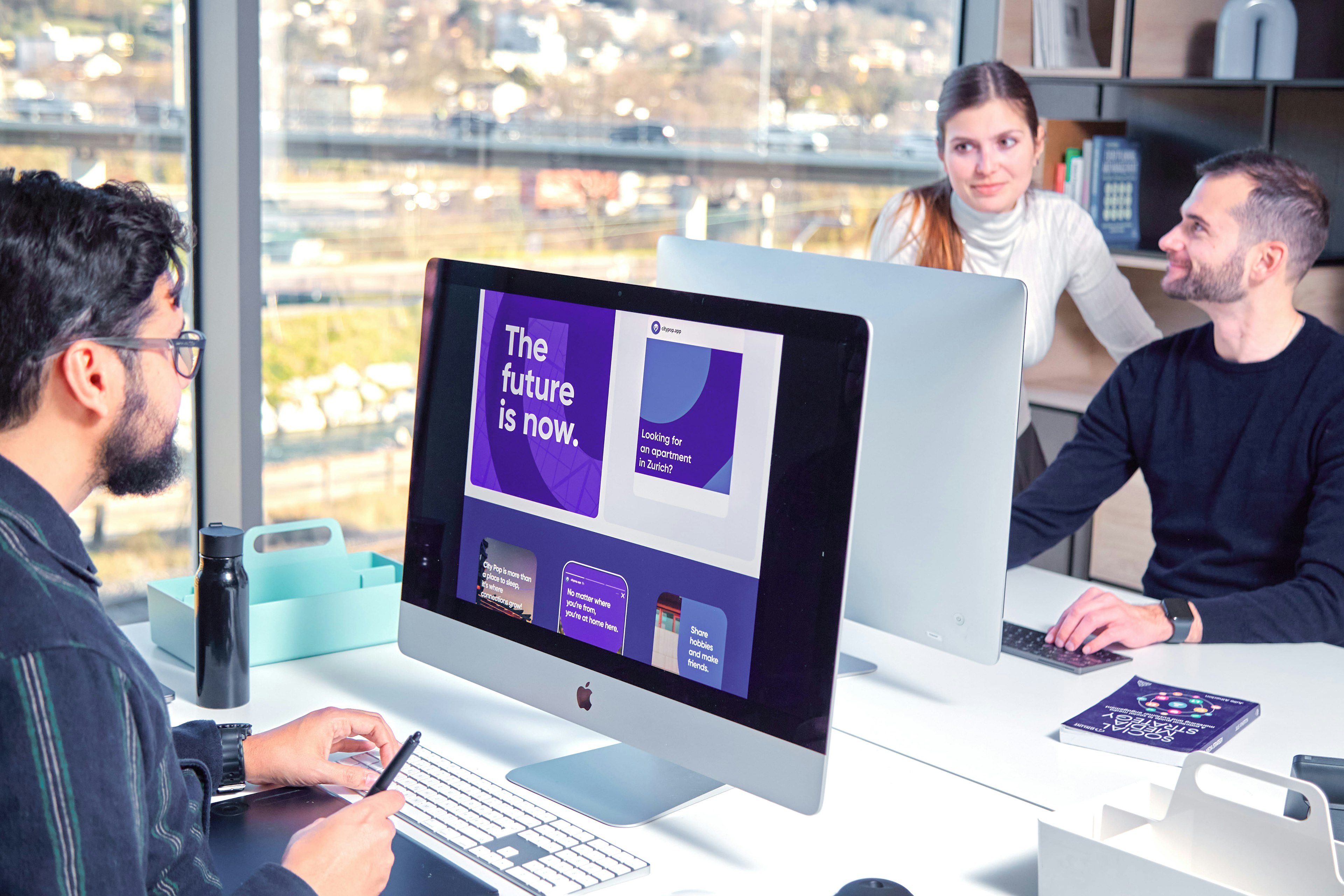 Three professionals in a modern office setting, collaborating around a large iMac displaying a vibrant purple presentation with the text 'The future is now.' The environment offers a view of an urban landscape through large windows