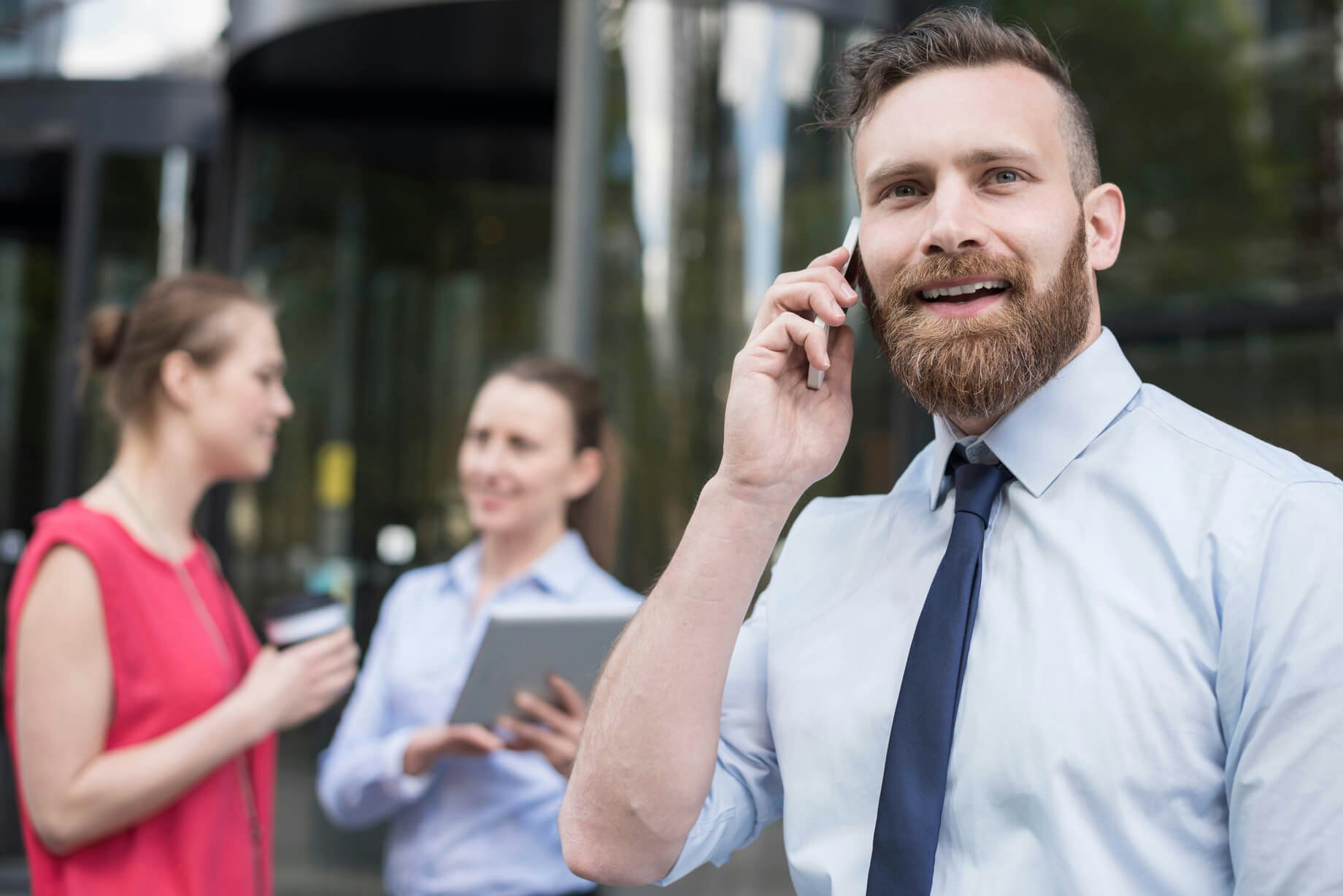 Uomo con barba al telefono sorride, sullo sfondo due colleghe parlano davanti a un edificio moderno.