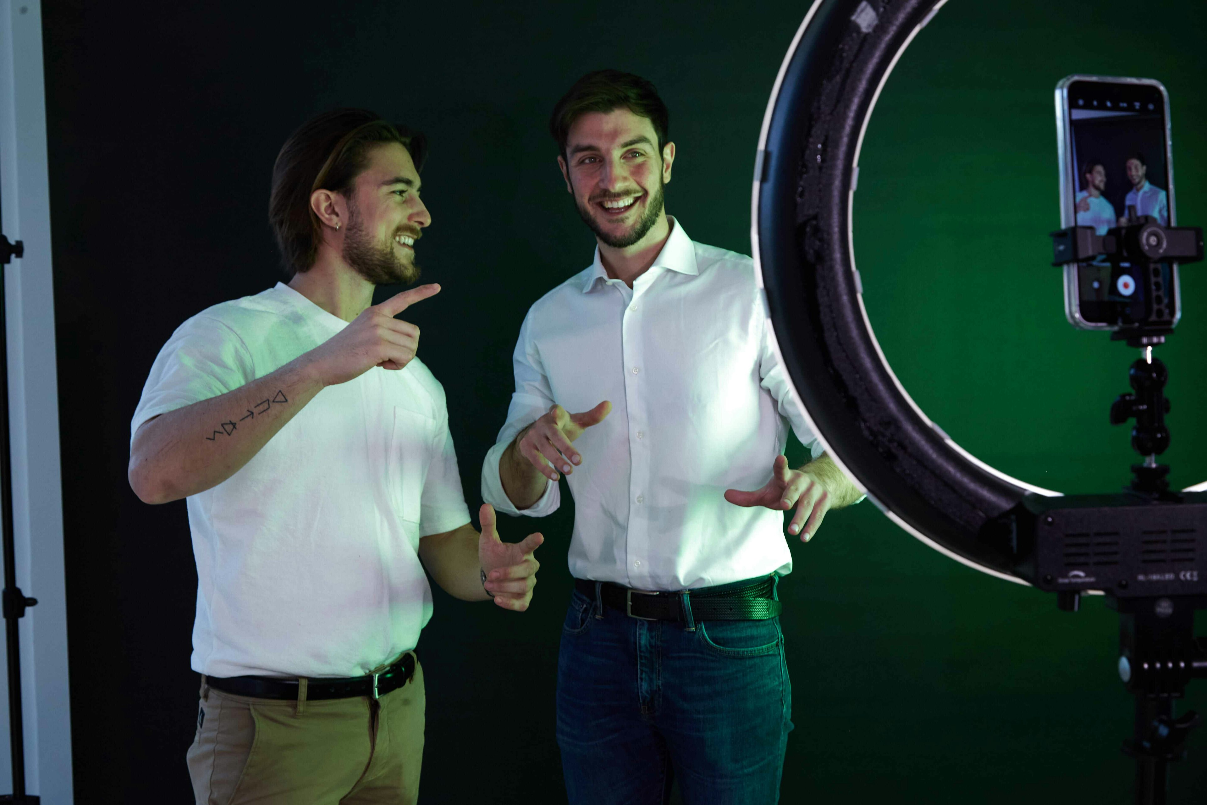 Two young men conversing in a studio with a ring light and camera setup, one in a white t-shirt and the other in a white shirt, both gesturing animatedly.