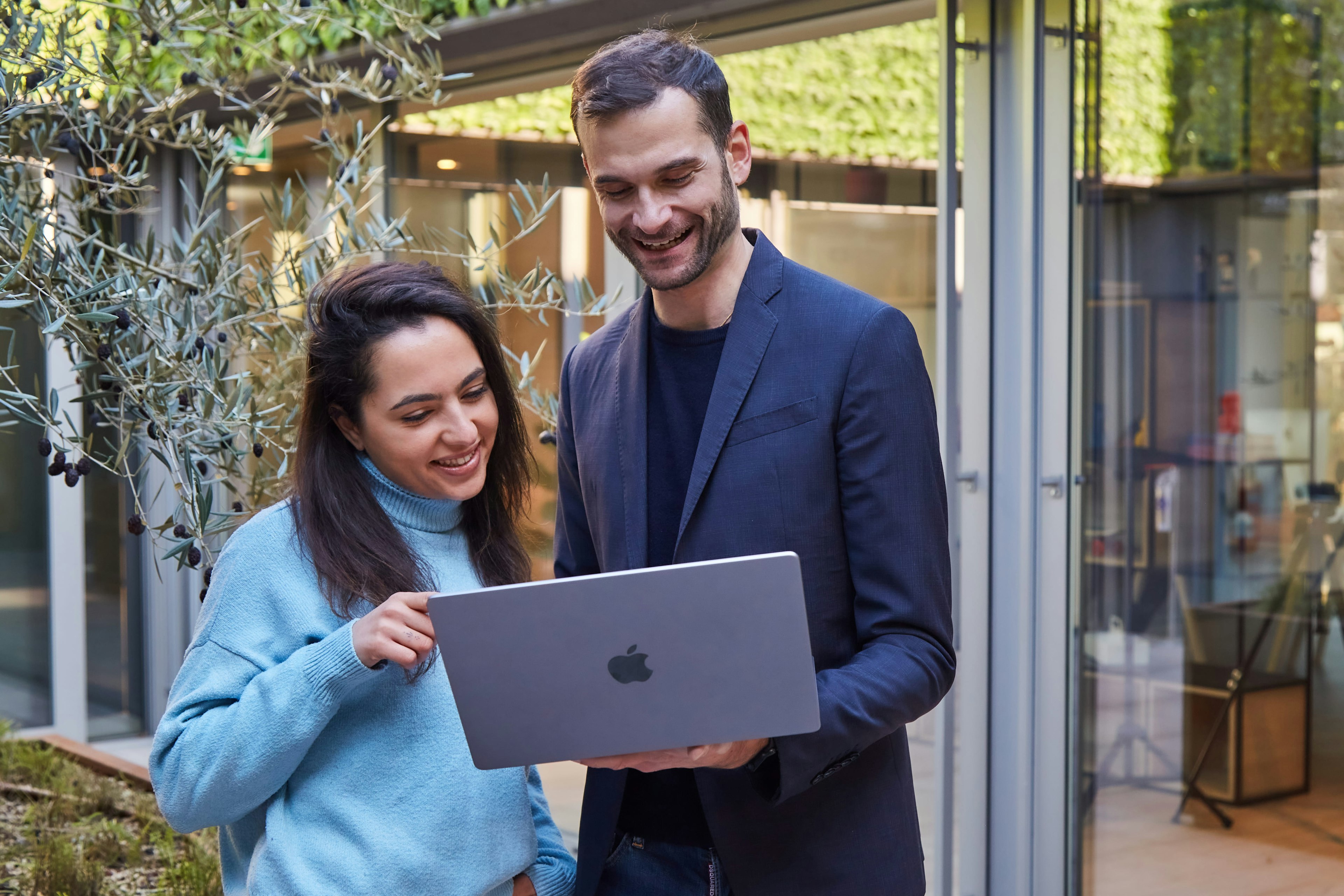 A man and a woman reviewing content on a laptop outdoors, surrounded by lush greenery and modern office buildings