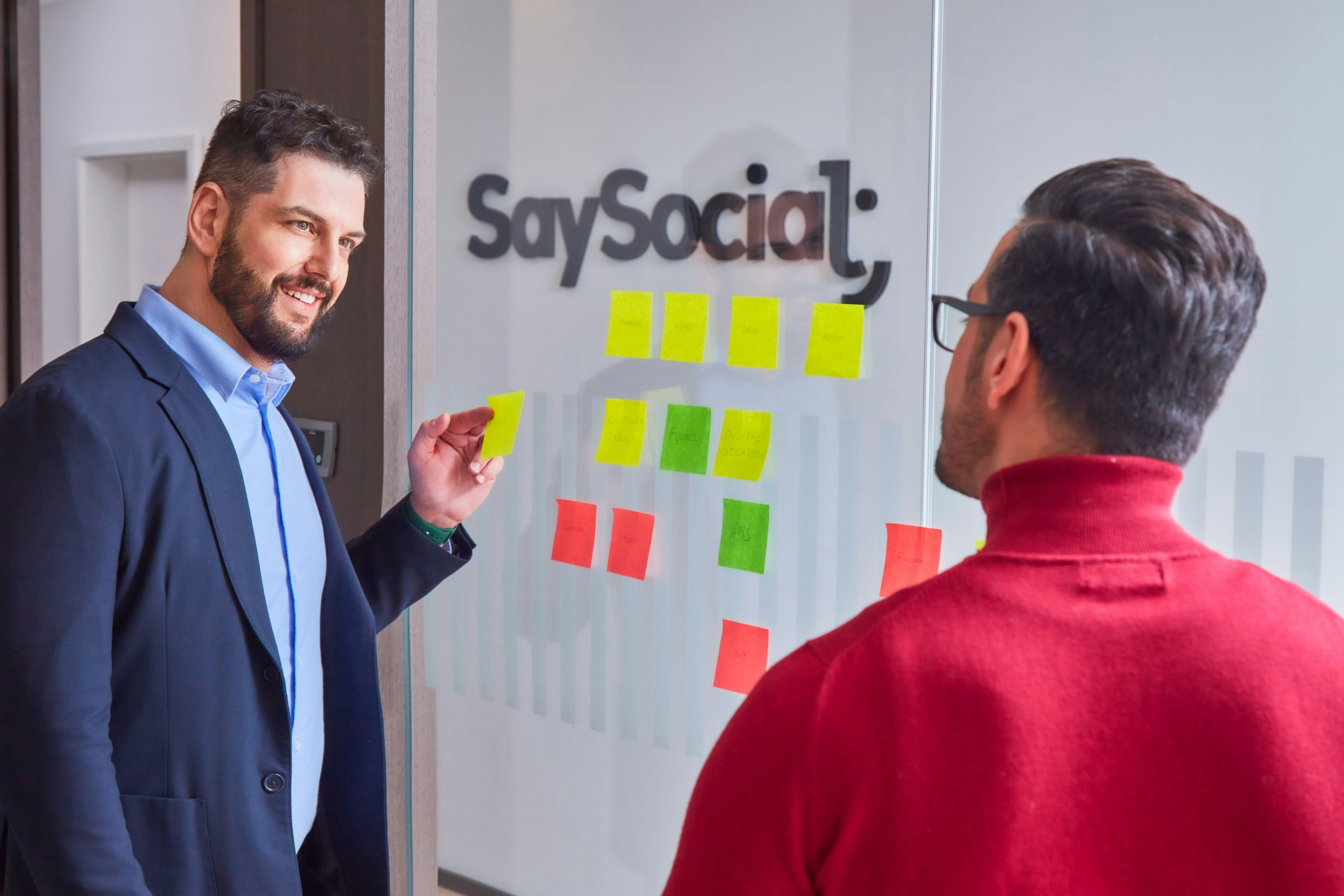 Two men engaging in a strategic discussion in an office setting, with one man pointing at sticky notes on a glass wall that feature terms like 'Audience,' 'Funnel,' and 'Ads,' under the logo 'SaySocial' in the background.