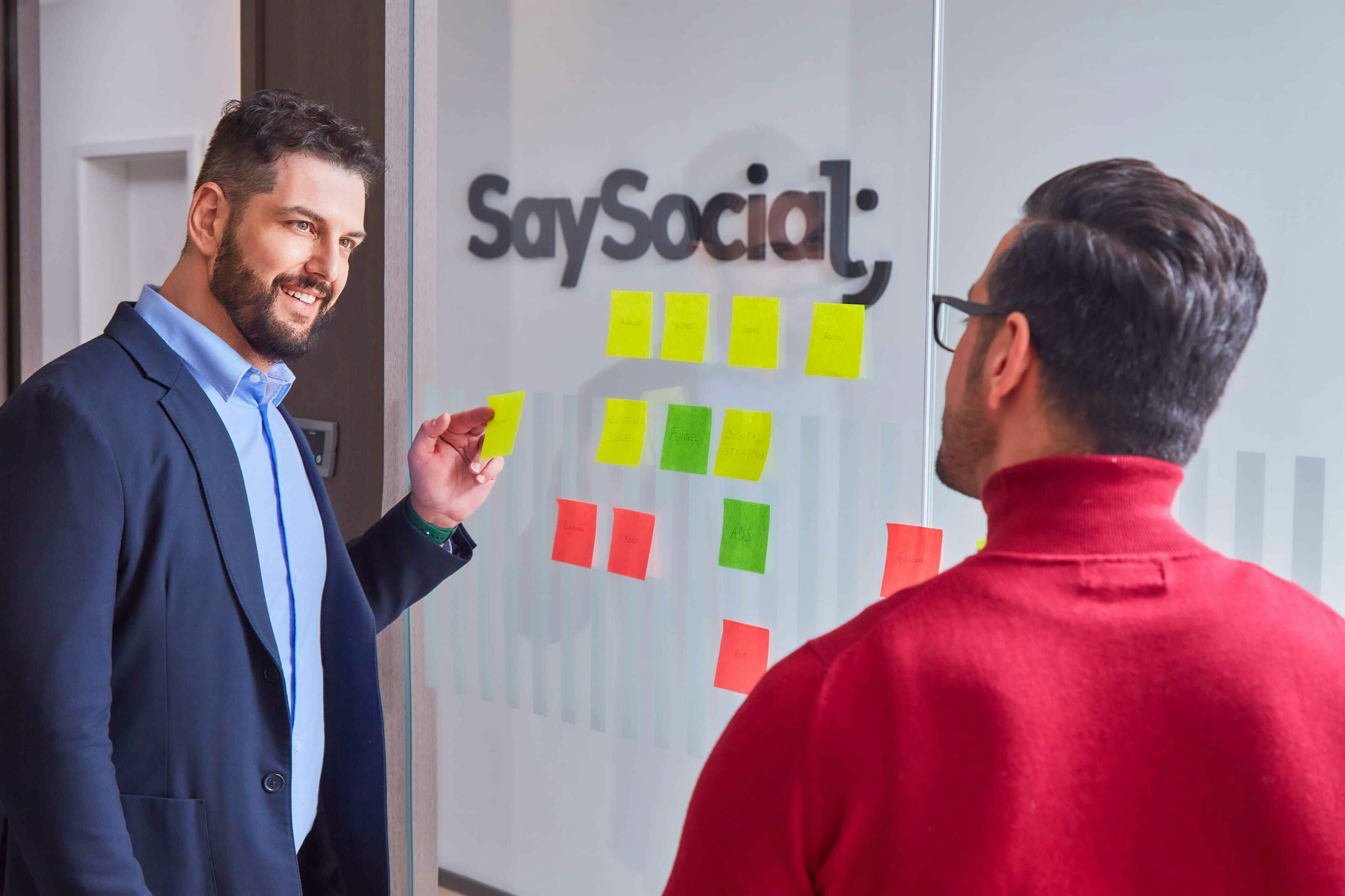 wo men engaging in a strategic discussion in an office setting, with one man pointing at sticky notes on a glass wall that feature terms like 'Audience,' 'Funnel,' and 'Ads,' under the logo 'SaySocial' in the background