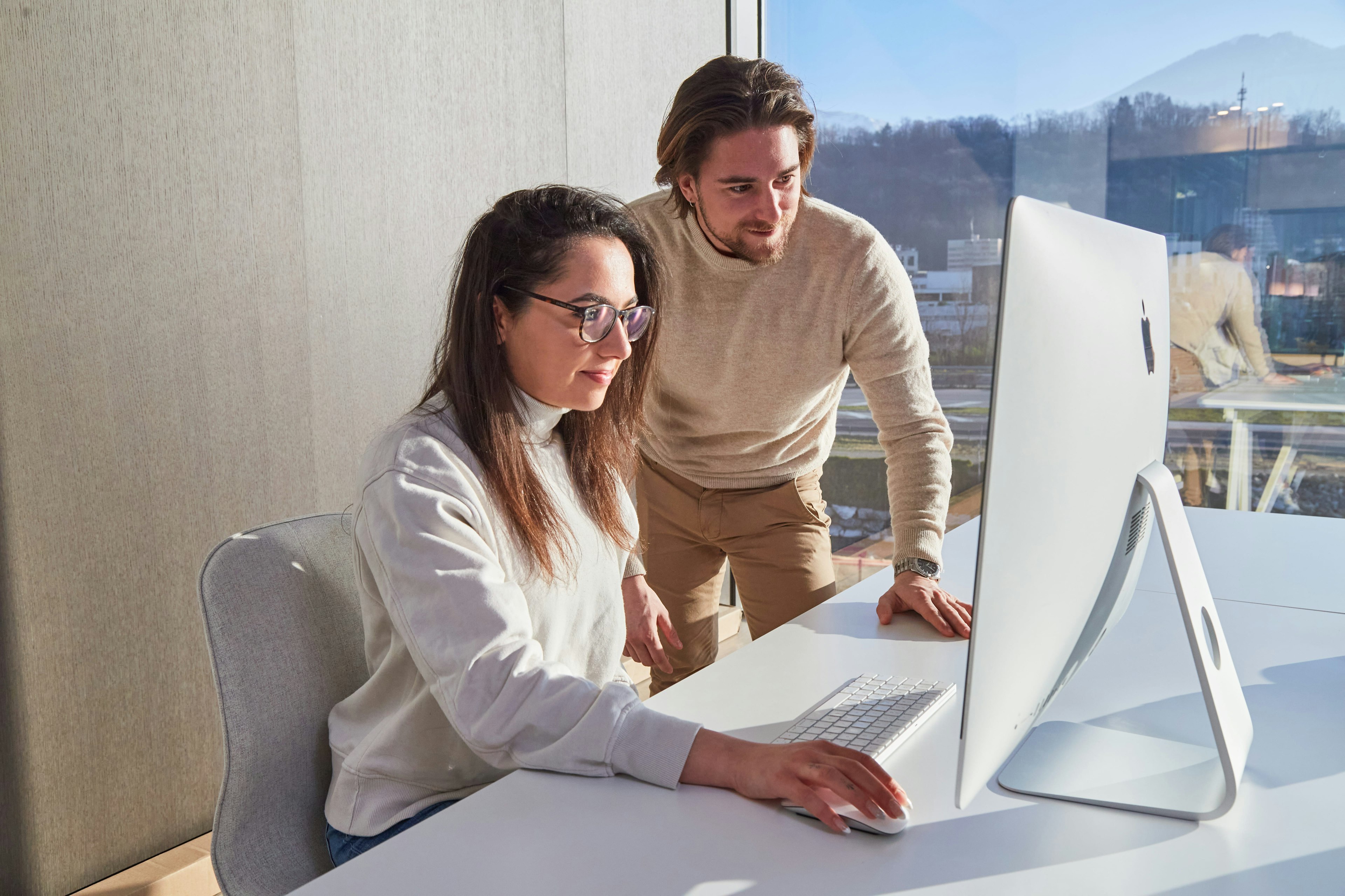 Two professionals, a man and a woman, collaboratively working at a computer in a bright office, focusing intently on the screen.