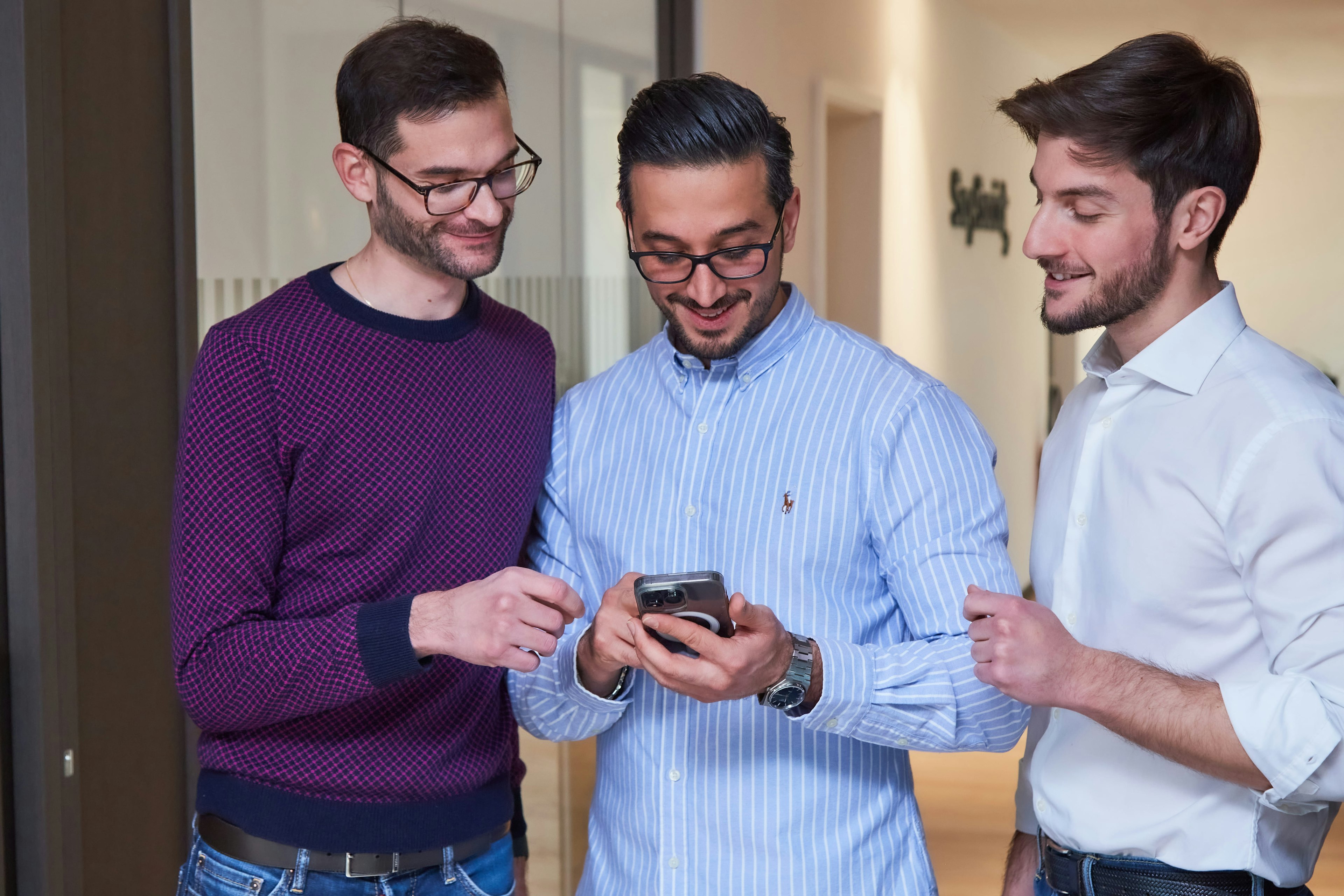 Three men standing together in an office environment, looking at a smartphone that one of them is holding. They are engaged in a discussion, possibly reviewing something on the device.