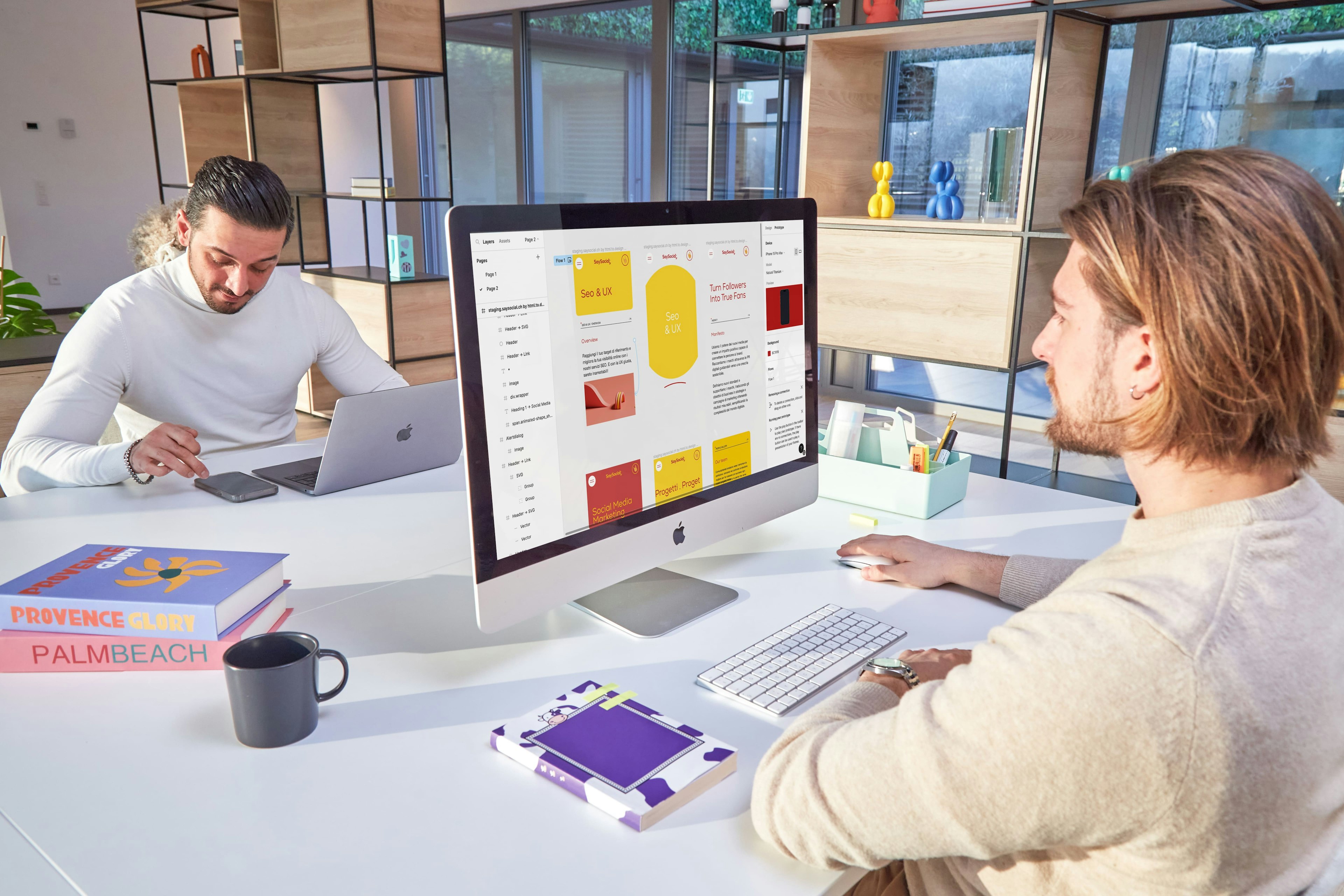 Two professionals collaborating in a bright, modern office, with one working on a laptop and the other reviewing data on a large monitor displaying colorful sticky notes and graphs.