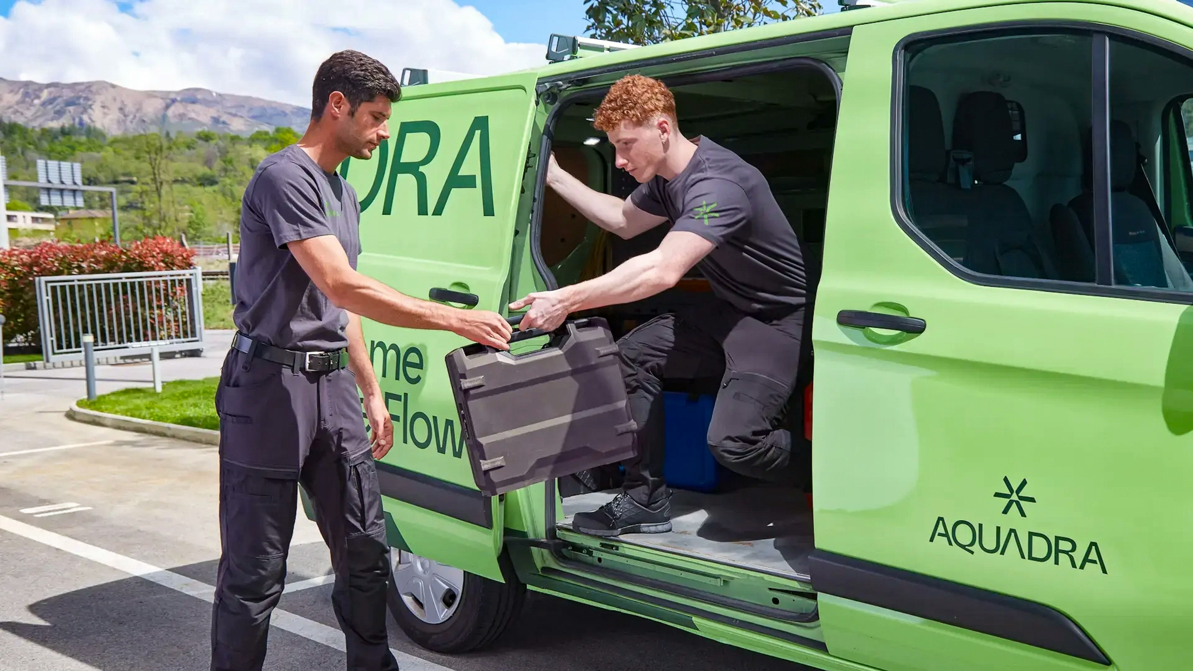 Two workers in black uniforms load a large equipment case into a bright green van labeled 'AQUADRA' in an outdoor parking area, with a mountain range in the background.