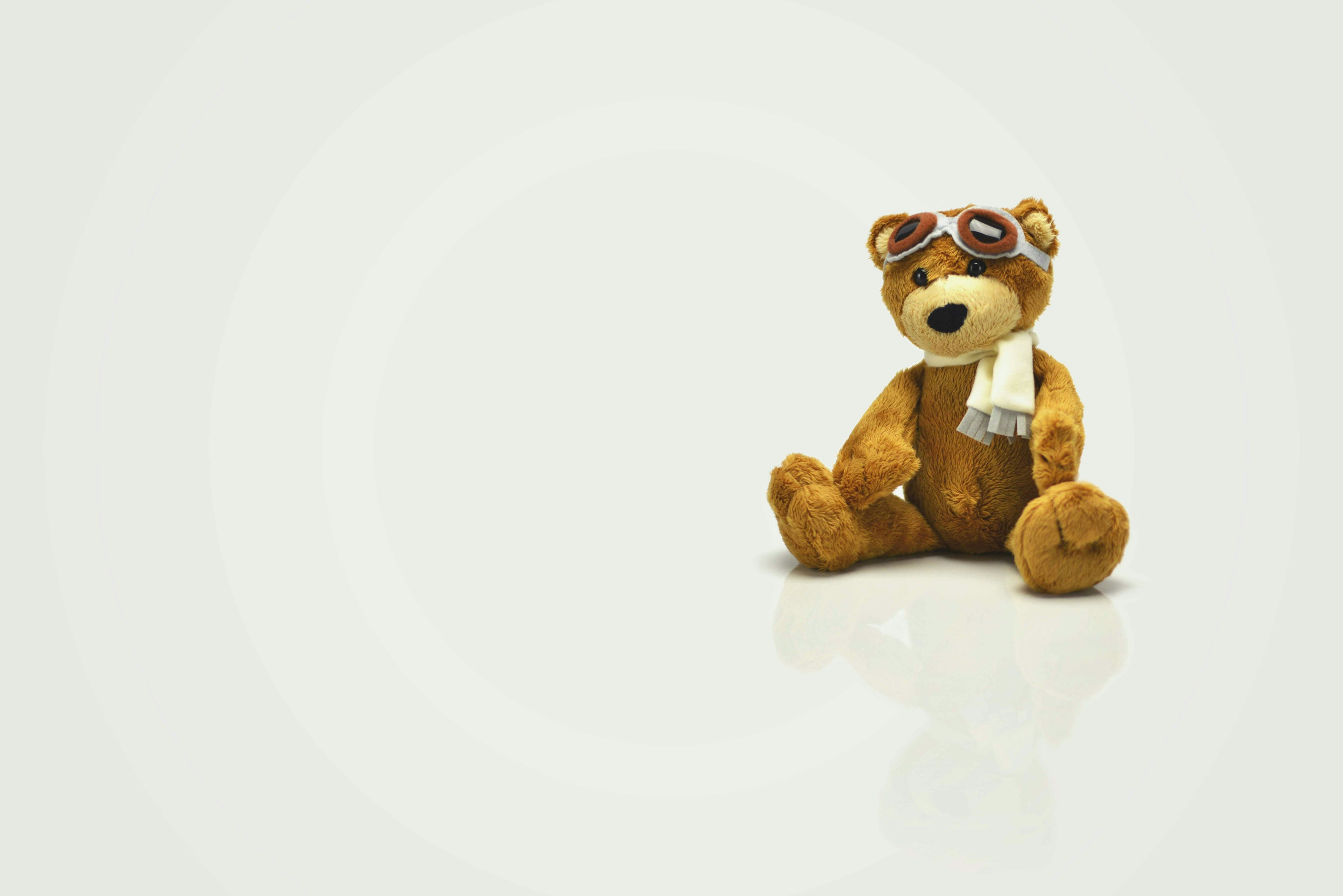A brown teddy bear wearing aviator goggles and a white scarf sits on a reflective surface against a light, minimalist background. The image emphasizes simplicity, nostalgia, and the charm of childhood.