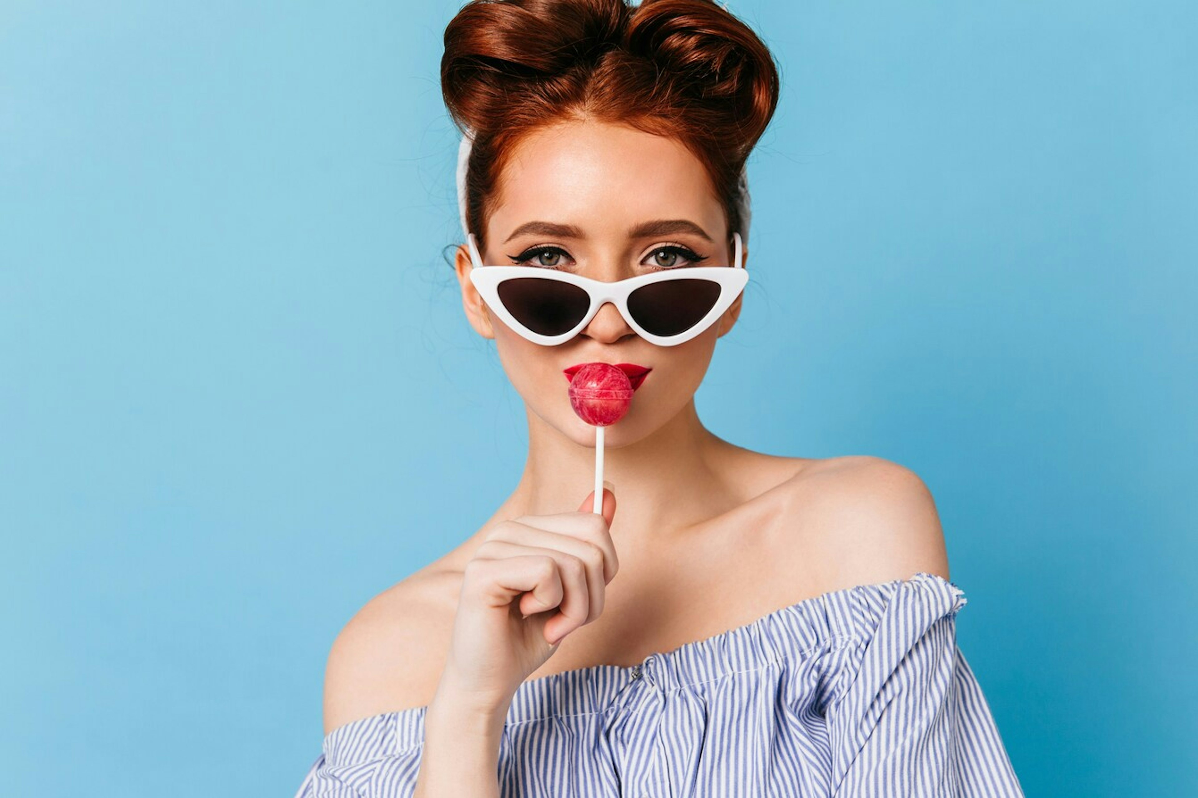 Young woman with red hair, wearing white cat-eye sunglasses, holds a red lollipop to her lips against a light blue background. Fashionable and playful vibe.