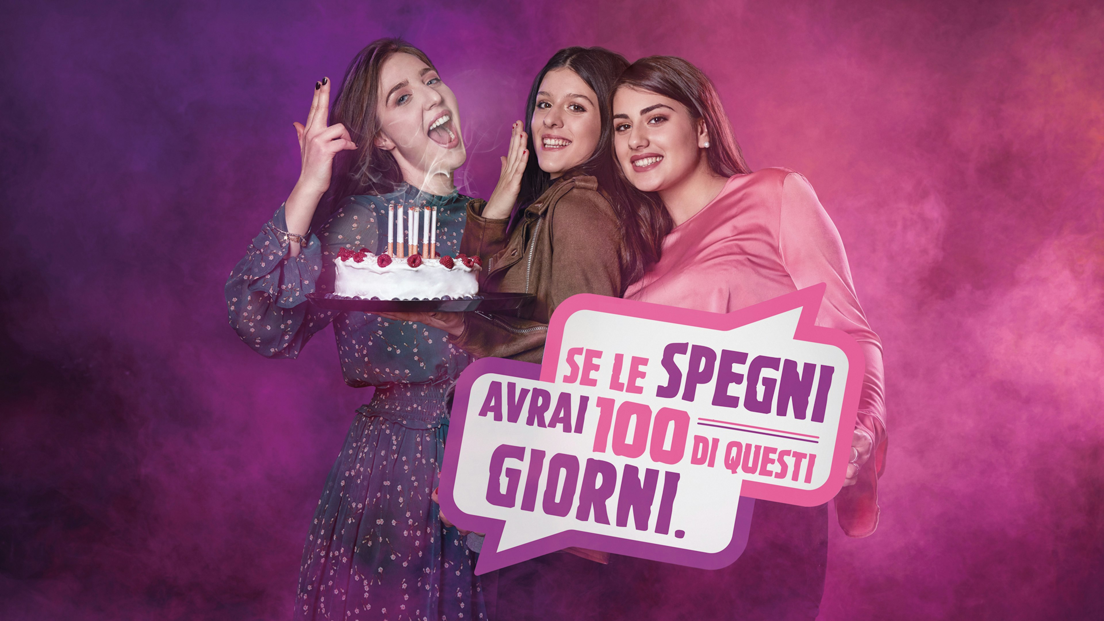Three young women hold a birthday cake with lit cigarettes as candles. A bold speech bubble reads, "Se le spegni, avrai 100 di questi giorni," conveying an anti-smoking message. The background is filled with pink and purple smoke.