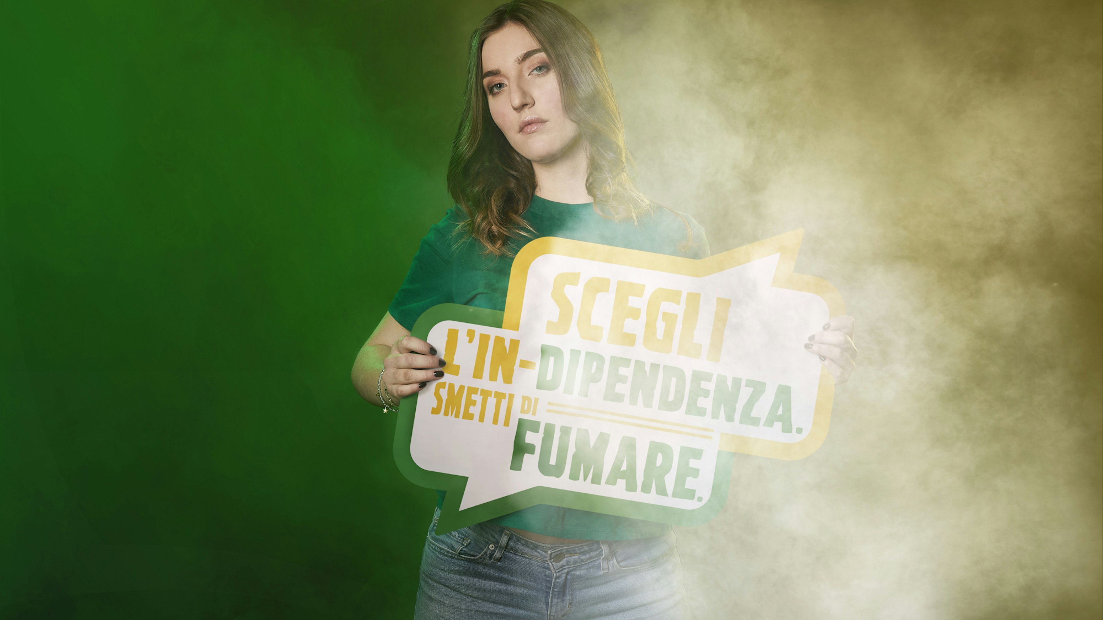 A young woman holds a sign reading "Scegli l'indipendenza. Smetti di fumare." She stands against a green and yellow smoky background, emphasizing the anti-smoking message.