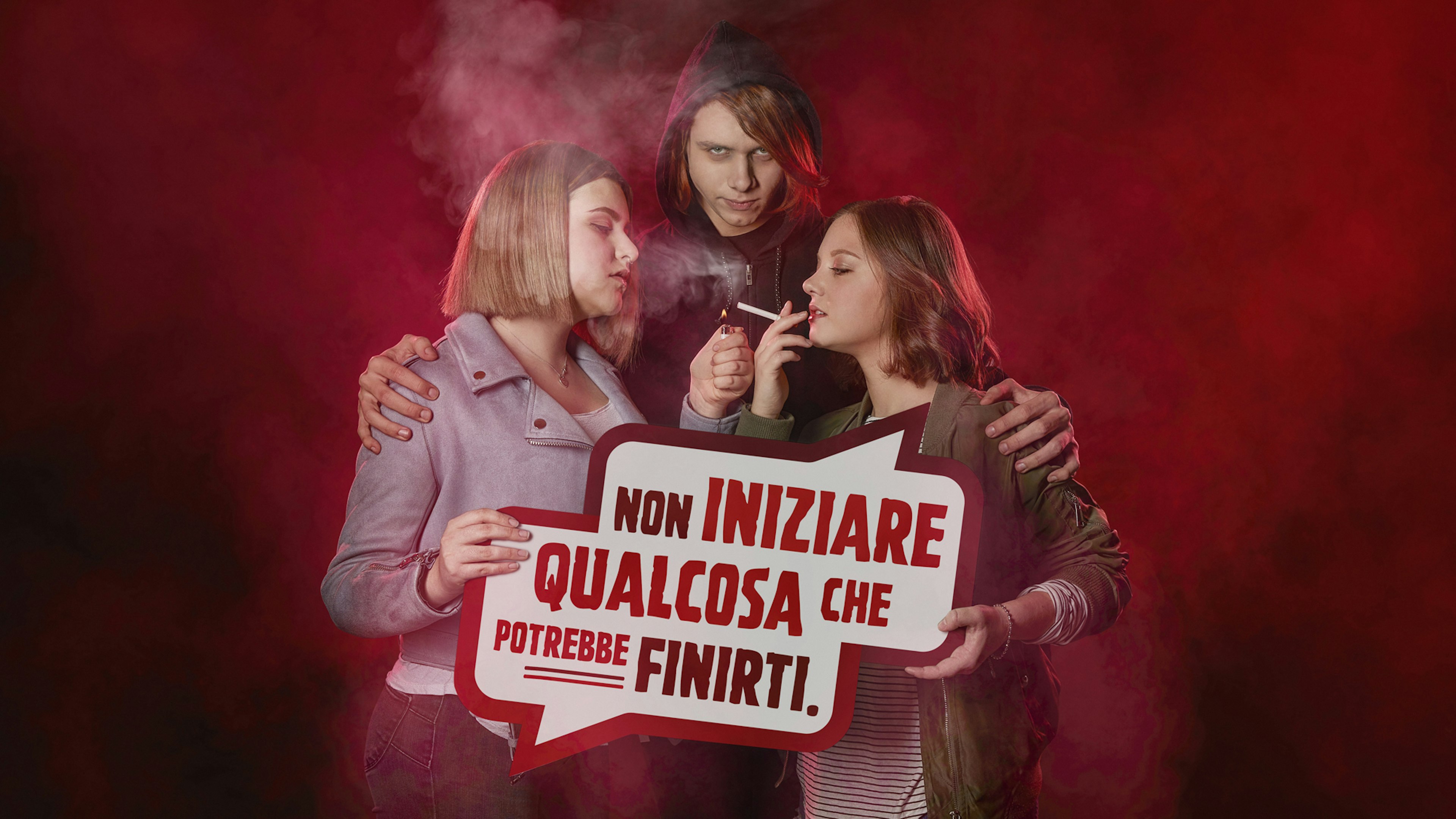 Three youths in a smoky red scene, two lighting cigarettes. A speech bubble reads, "Non iniziare qualcosa che potrebbe finirti," conveying an anti-smoking message.